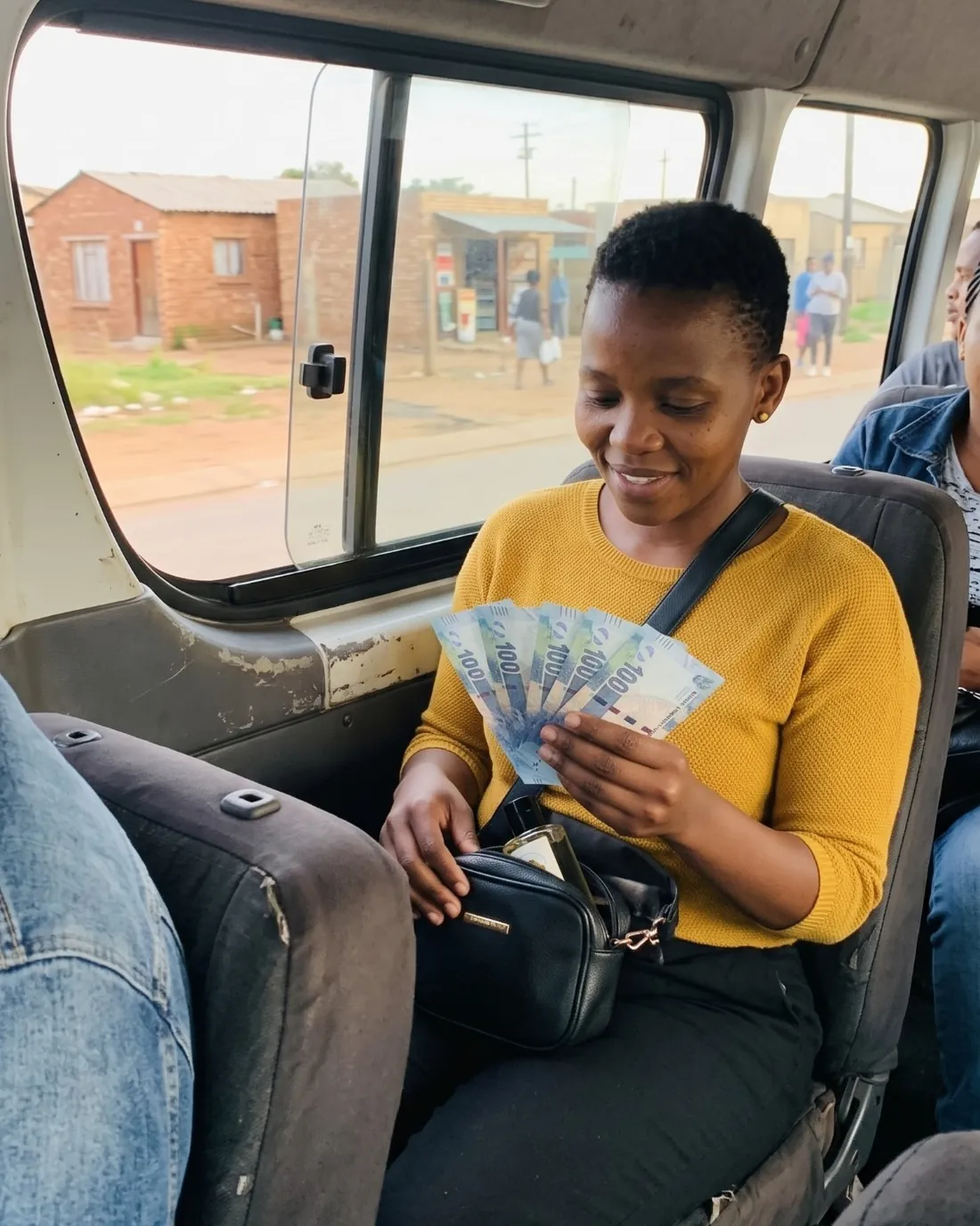 A woman in a mustard top in a minibus taxi, smiling at the cash in her hand