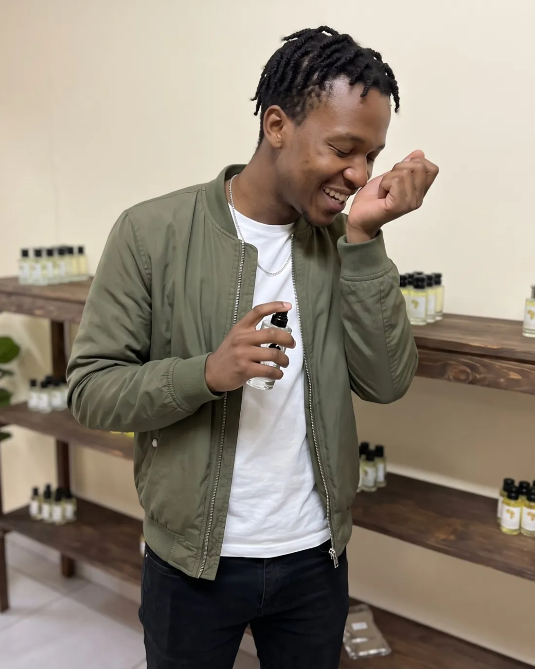 A young man in an olive jacket smelling his wrist after one spray, grinning