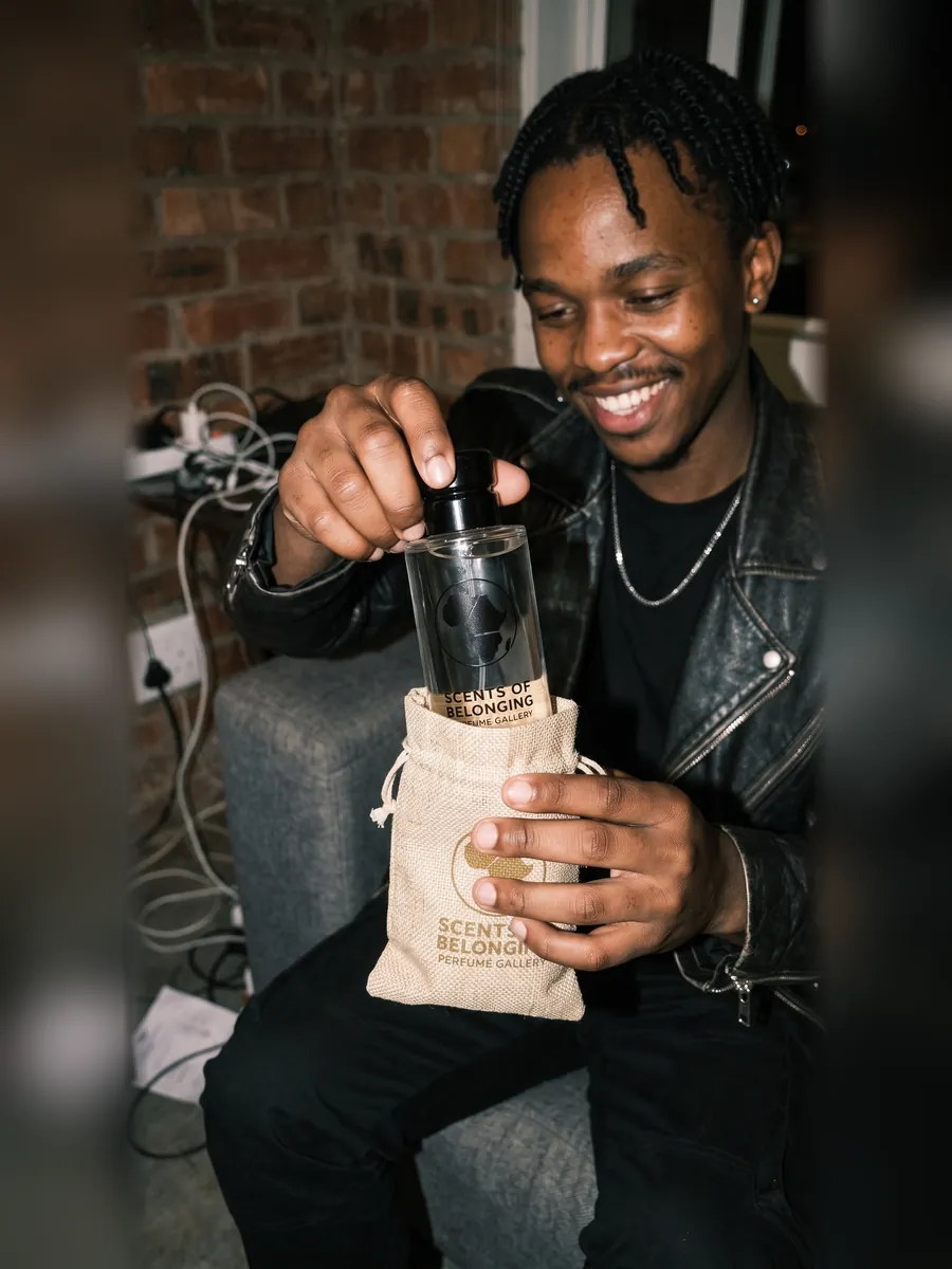 A young man in a black leather jacket smiling as he lifts his Scents of Belonging bottle out of its jute pouch